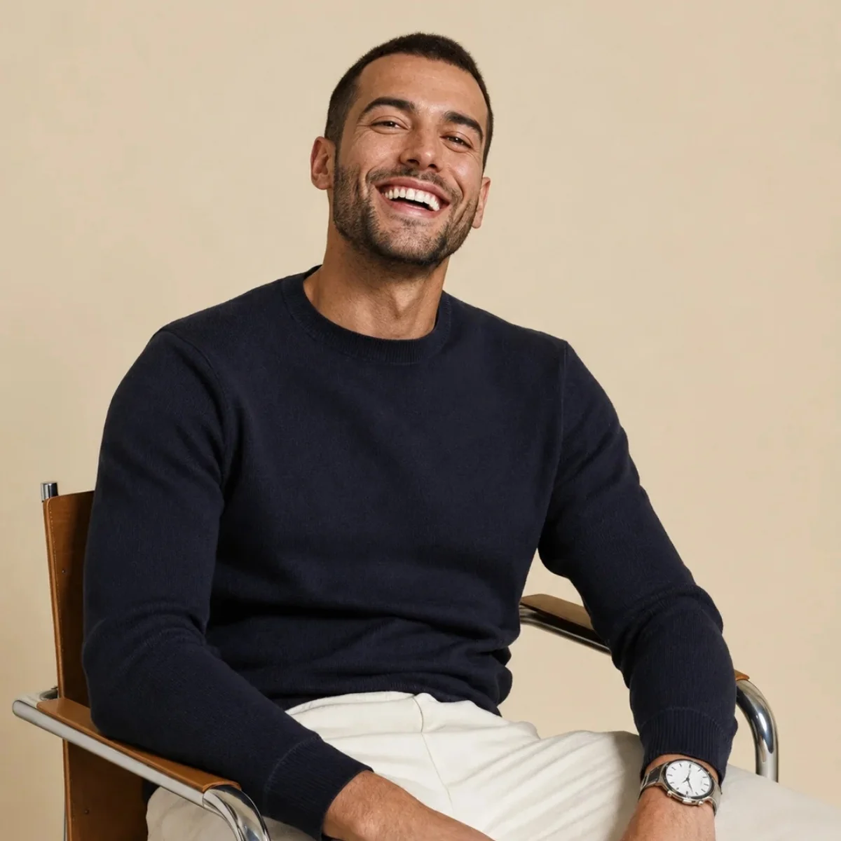 Design sans titre (11) Homme d'une trentaine d'années souriant, pull cachemire marine et pantalon crème, assis sur une chaise cantilever, fond beige